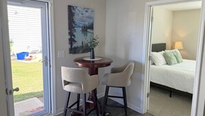 A room with a bed, a small table with chairs, a painting on the wall, and a door leading outside.