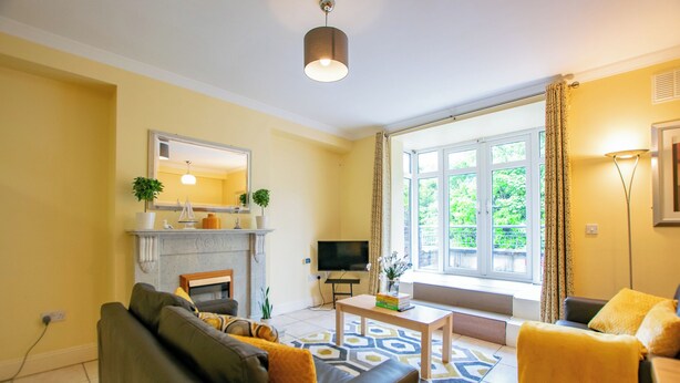 Ett vardagsrum med en soffa, ett soffbord, en öppen spis, en tv och ett stort fönster med gardiner.