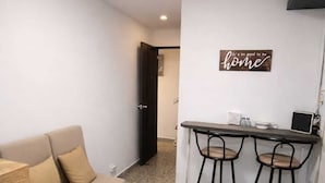 Un espacio habitable compacto con sofá, una mesita redonda, una zona de cocina con un taburete y un cartel en la pared.