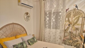 Ein Zimmer mit einem Bett, einer Wand-Klimaanlage, einem dekorativen Spiegel mit tropischem Motiv und einem Fenster mit leichtem Vorhang.