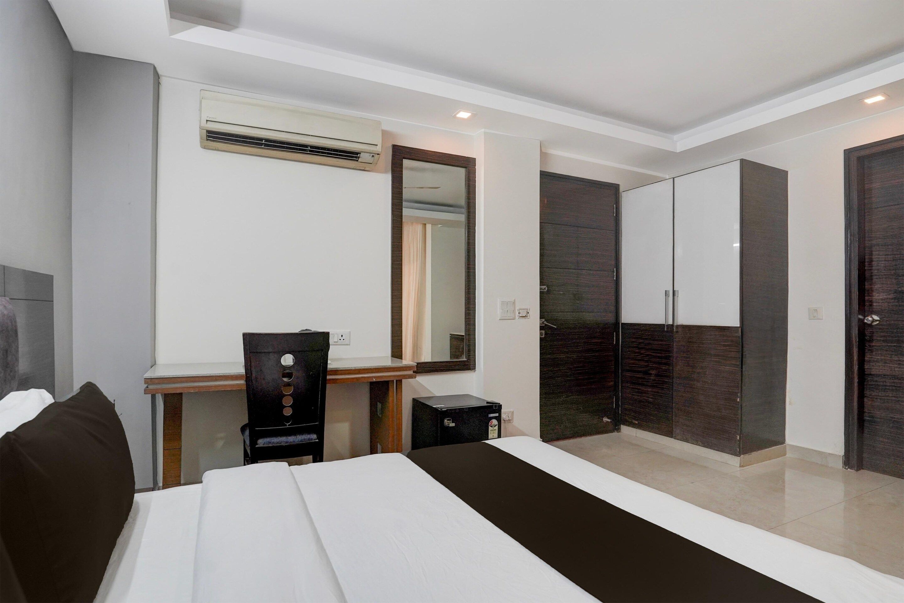 A hotel room with a bed, a desk, a chair, a mirror, a wardrobe, and an air conditioning unit.