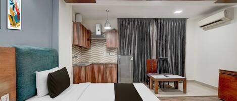 A hotel room with a bed, a desk, a chair, a small table, an air conditioning unit, a refrigerator, a wall-mounted telephone, and a decorative wall piece.