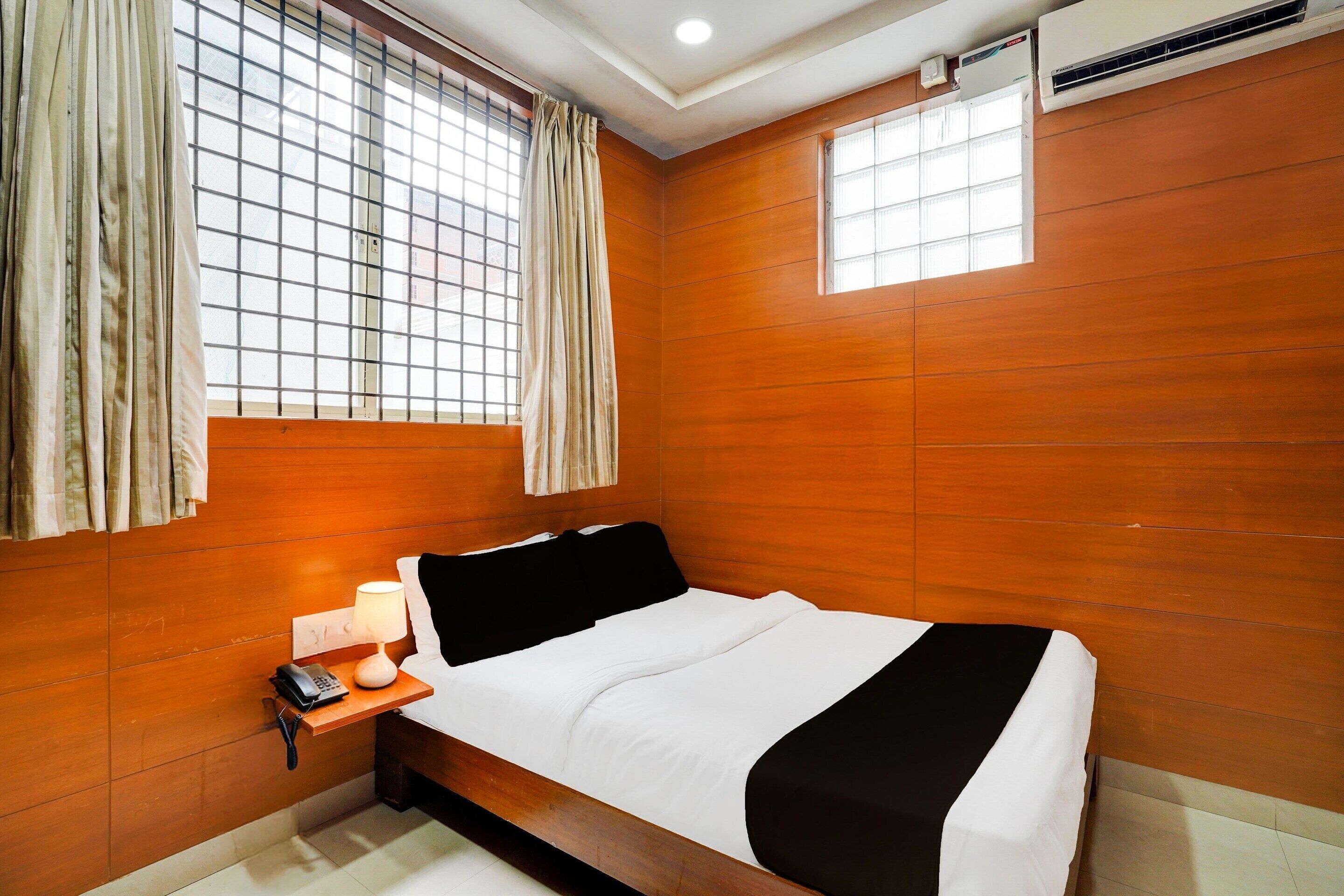 A hotel room with a wooden wall, a bed with white bedding, a bedside table with a lamp, a phone, and a window with curtains.