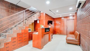 A reception area with a wooden reception desk, a leather sofa, a staircase with a glass railing, and a wall-mounted air conditioning unit. - Deluxe Collection O Kalyan Nagar Bangalore (Bengaluru)