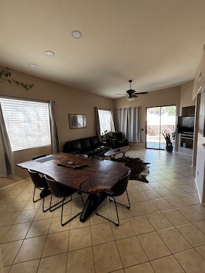 A spacious living area with a large wooden dining table, a black leather sofa, a ceiling fan, and a TV in the background. -  Saguaro West Retreat, Fire Pit, Desert Views & Peaceful Stay! Animal friendly (Tucson)