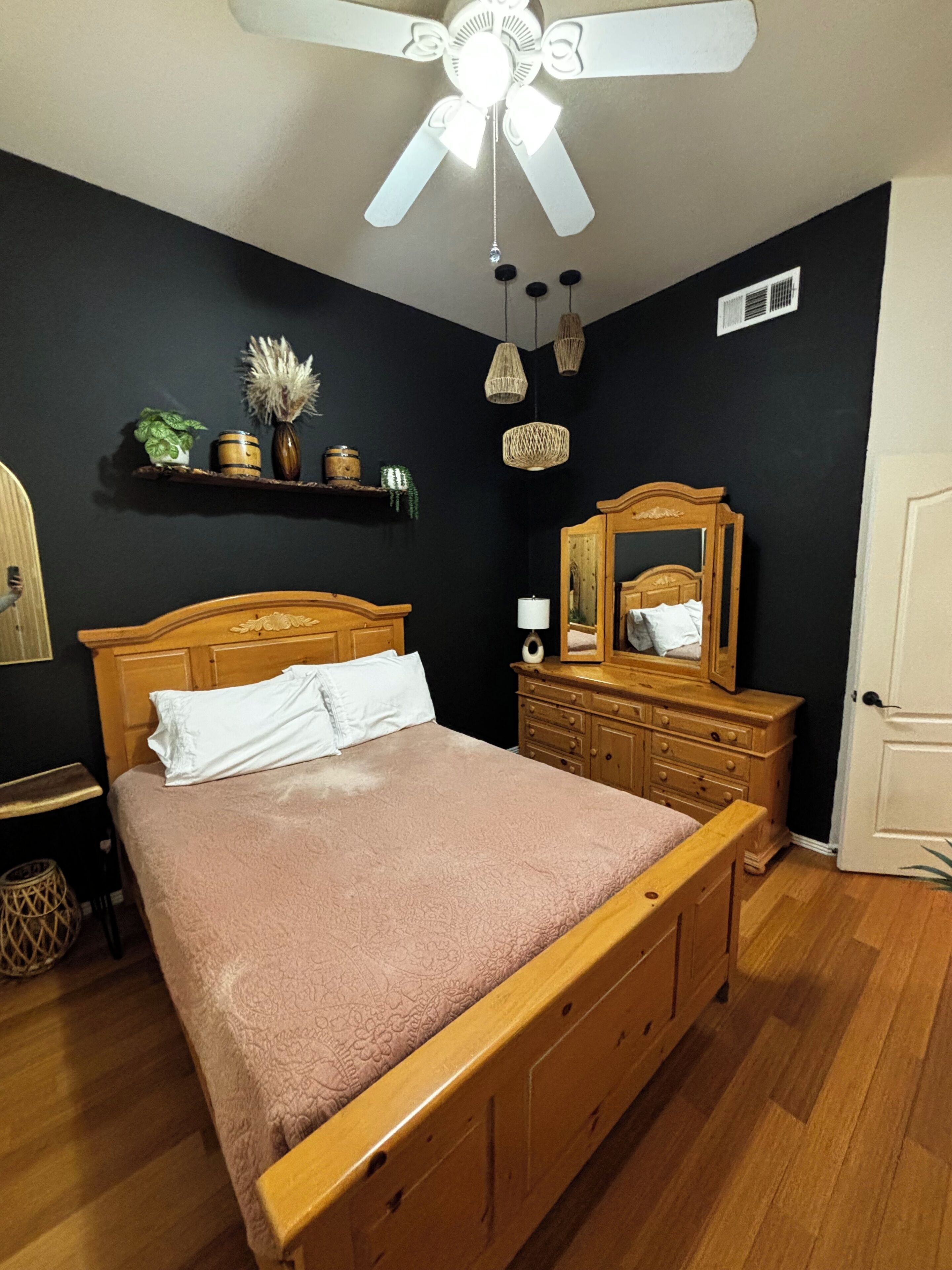 Una camera da letto con un letto di legno, una ventola a soffitto, una cassettiera, uno specchio e una mensola con oggetti decorativi.