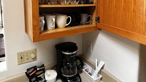 Ett kök med ett träskåp som innehåller glas och muggar, en kaffebryggare och kaffekapslar på bänkskivan.