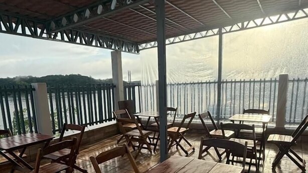 An outdoor patio with wooden tables and chairs, a metal railing, and a view of the water. - Hotel beira rio OPK (Oiapoque)