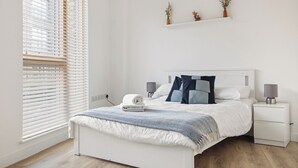 Een slaapkamer met een bed, nachtkastjes, een venster met zonwering en een plank met decoratieve objecten.