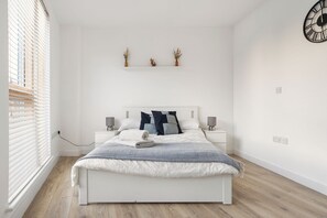 Une chambre à coucher moderne avec un lit blanc, des tables de chevet, une horloge et une fenêtre avec des stores.