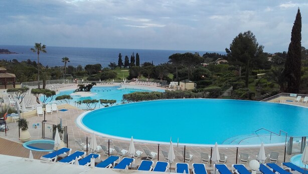 Ein großer Swimmingpool mit blauem Wasser, umgeben von Liegestühlen und Sonnenschirmen, mit Blick auf das Meer und Bäume im Hintergrund.