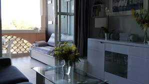 Ein modernes Wohnzimmer mit Glastisch, einer Couch, einem Bett mit Blick ins Freie, einer Küchenzeile mit weißem Schrank und einem gerahmten Bild an der Wand.
