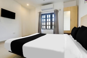 A hotel room with a bed, a flat-screen TV mounted on the wall, an air conditioning unit, a window with curtains, and a wardrobe.