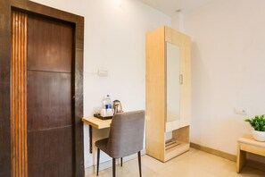 A room with a wooden door, a desk with a chair, a mirror, a small table with a plant, and a light switch on the wall.