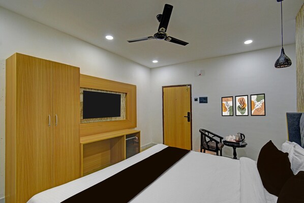 A hotel room with a bed, a TV, a ceiling fan, a desk with a chair, and artwork on the wall.