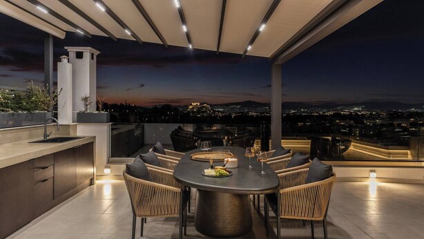 Una terraza en la azotea con una mesa redonda, sillas, una zona de bar y vistas a la ciudad al atardecer.