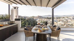 Un área de comedor en la azotea con una mesa preparada para cuatro personas, rodeada de sillas de mimbre, con vistas a un paisaje urbano que incluye edificios y una colina.