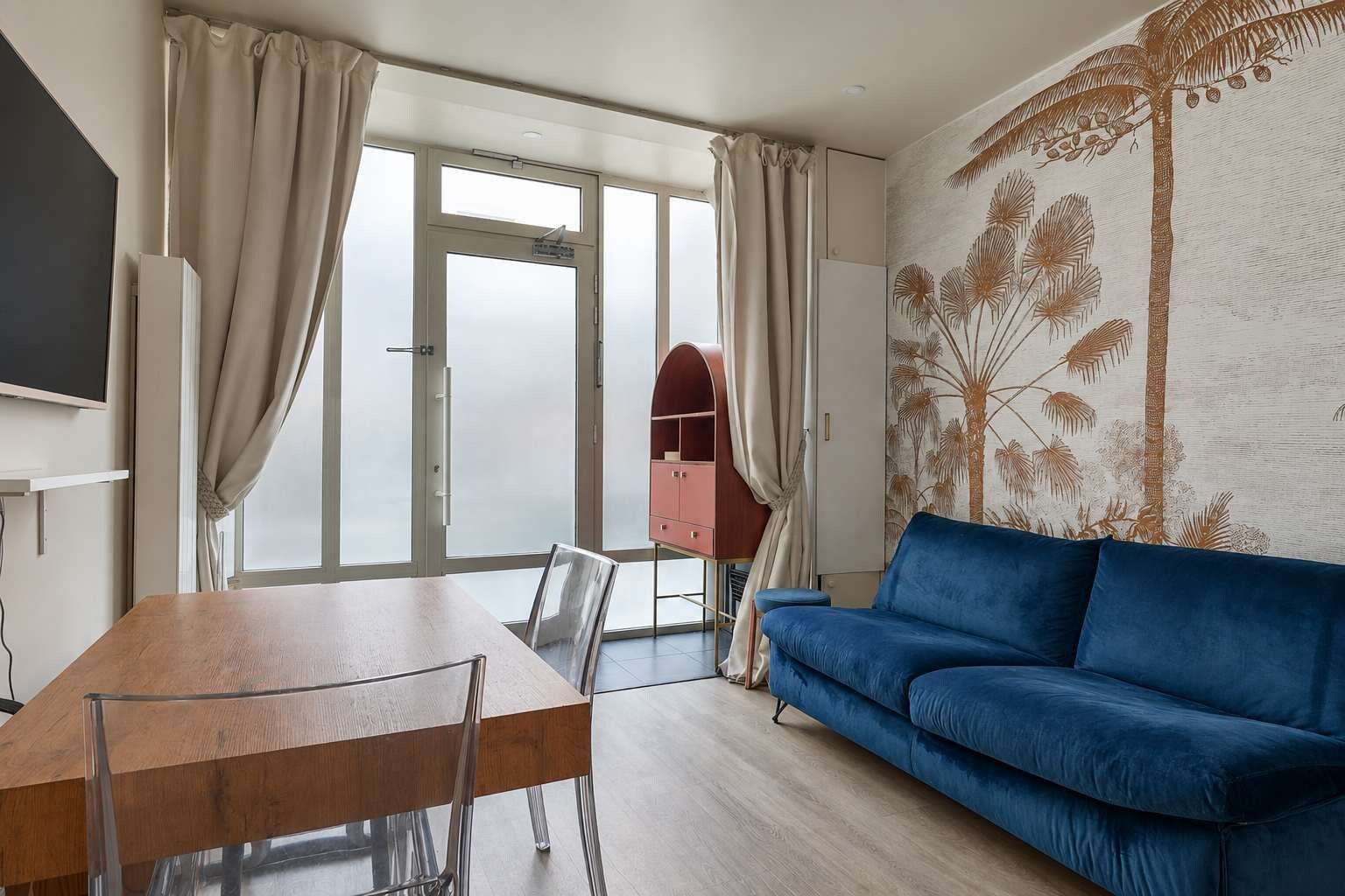 A modern living room with a wooden dining table, a blue sofa, and a large window with curtains.