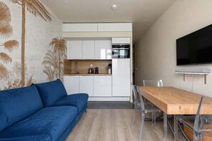 A modern living space with a sofa, a wooden dining table, clear chairs, a kitchen area with cabinets, and a flat-screen TV mounted on the wall. - Lumineux cocon pour 3 · Canal Saint-Martin (Paris)