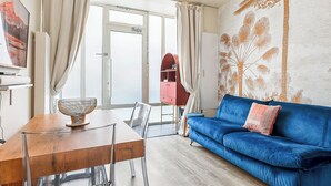 A modern living room with a blue sofa, a dining table with chairs, and a large window with curtains. - Cosy & lumineux – 3 pers – Paris centre (Paris)