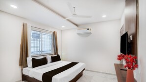 Een slaapkamer met een bed, een plafondventilator, een airconditioning, een raam met gordijnen, een televisie en een vaas met bloemen.