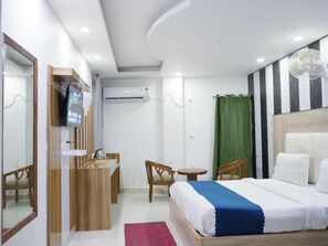 Ett hotellrum med en säng, ett skrivbord, en stol, ett litet bord, en spegel och en fläkt.