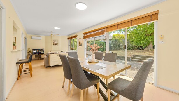 Un espace repas moderne avec une table en bois et des chaises grises, une cuisine en arrière-plan et une vue sur la verdure à l’extérieur.