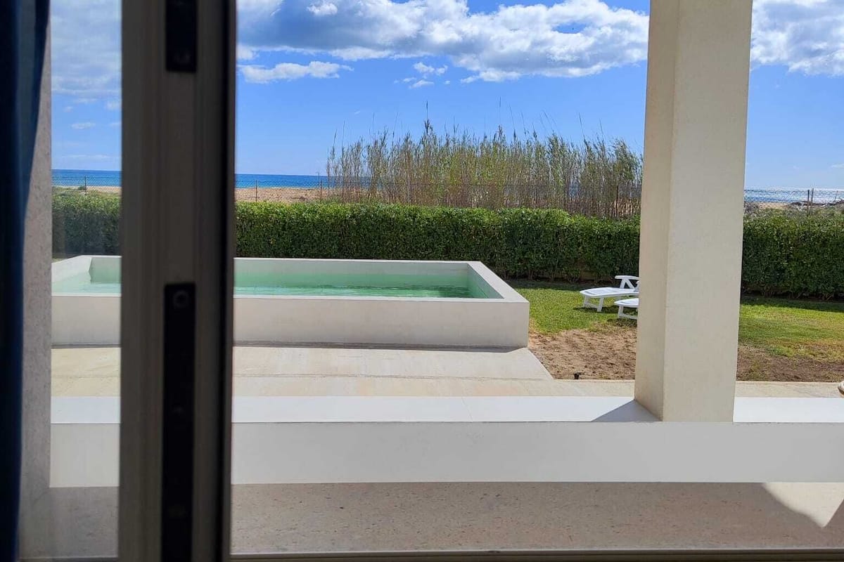A view through a window of a pool area with a clear sky, a grassy lawn, and a hedge.