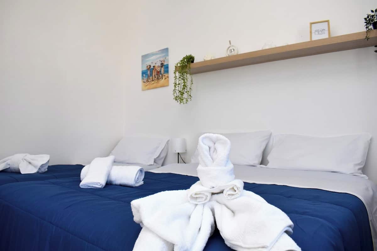 A hotel room with a bed, pillows, a bedside table, a wall-mounted shelf with decorative items, and a painting.