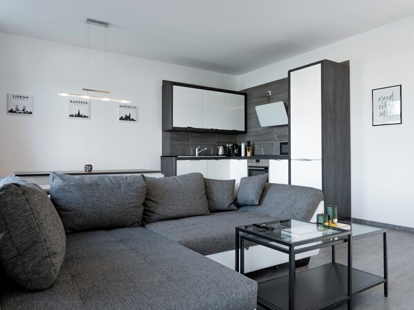 A modern living room with a sectional sofa, a glass coffee table, and a minimalist kitchen in the background. - Central apartment in Chemnitz + parking space - modern & well connected (Chemnitz)