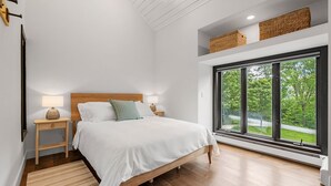 Ein Schlafzimmer mit großem Fenster, einem Bett mit weißer Bettwäsche, einem Holz-Kopfteil und zwei Nachttischen mit Lampen.
