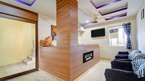 A hotel reception area with a wooden reception desk, a flat-screen TV mounted on the wall, a set of stairs leading to a room, and a seating area with patterned cushions. - Deluxe G K Building (Thindukkal)
