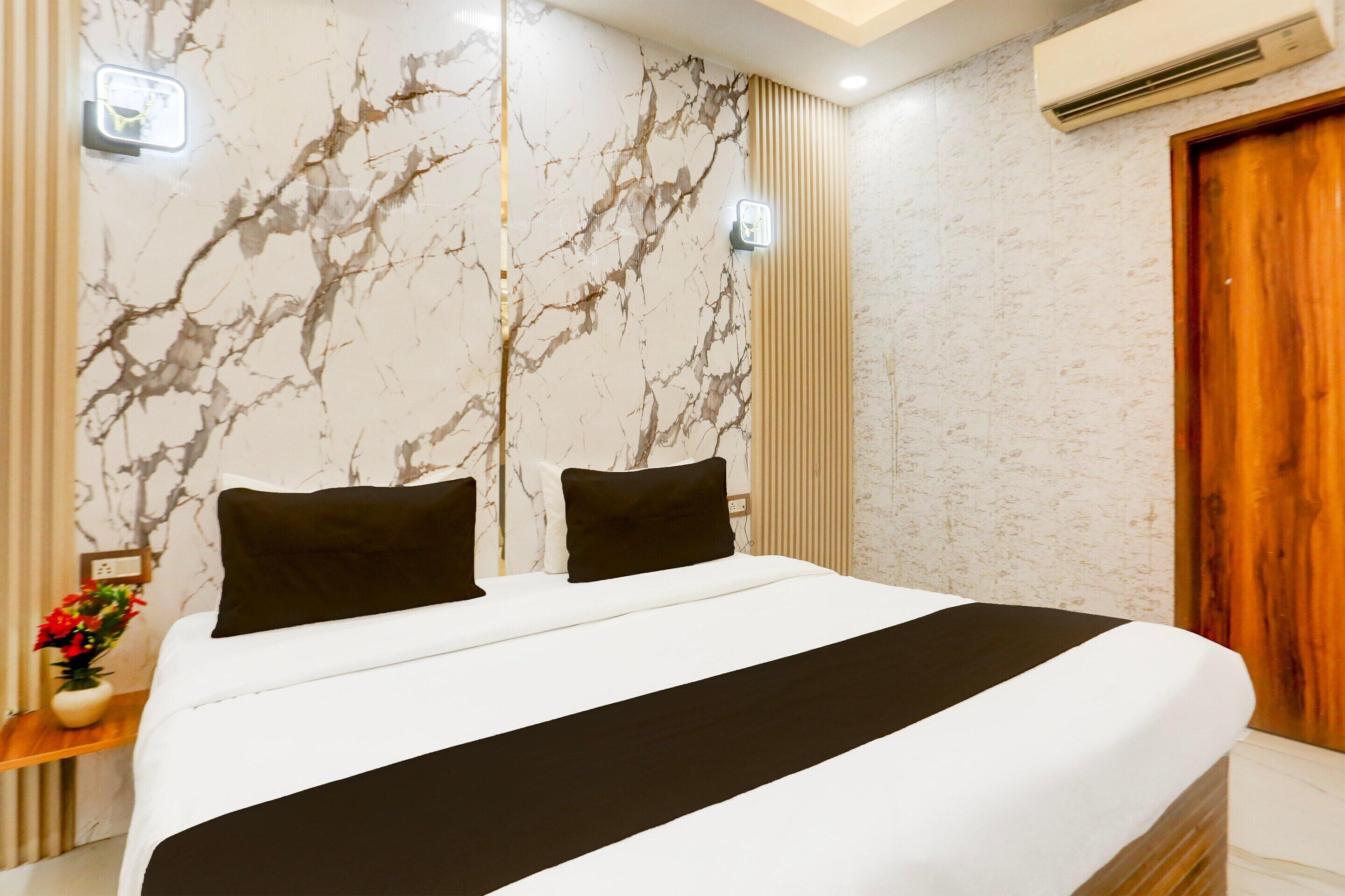 A hotel room with a marble-patterned wall, a wooden door, a bed with black and white bedding, and a nightstand with a vase of flowers.