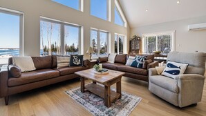 A spacious living room with a large window, a brown leather sofa, a wooden coffee table, and a beige armchair.