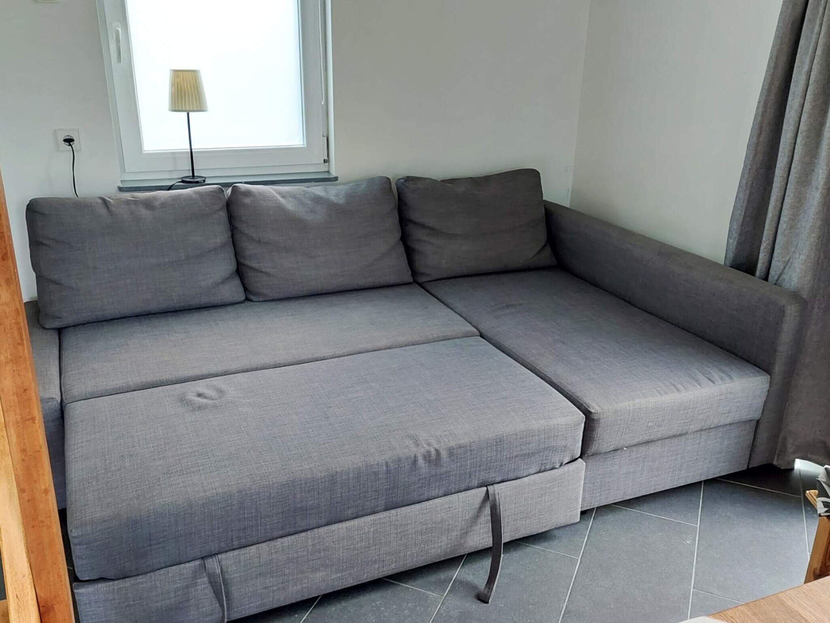 A grey sectional sofa with a pull-out bed, a lamp, and a window with curtains.