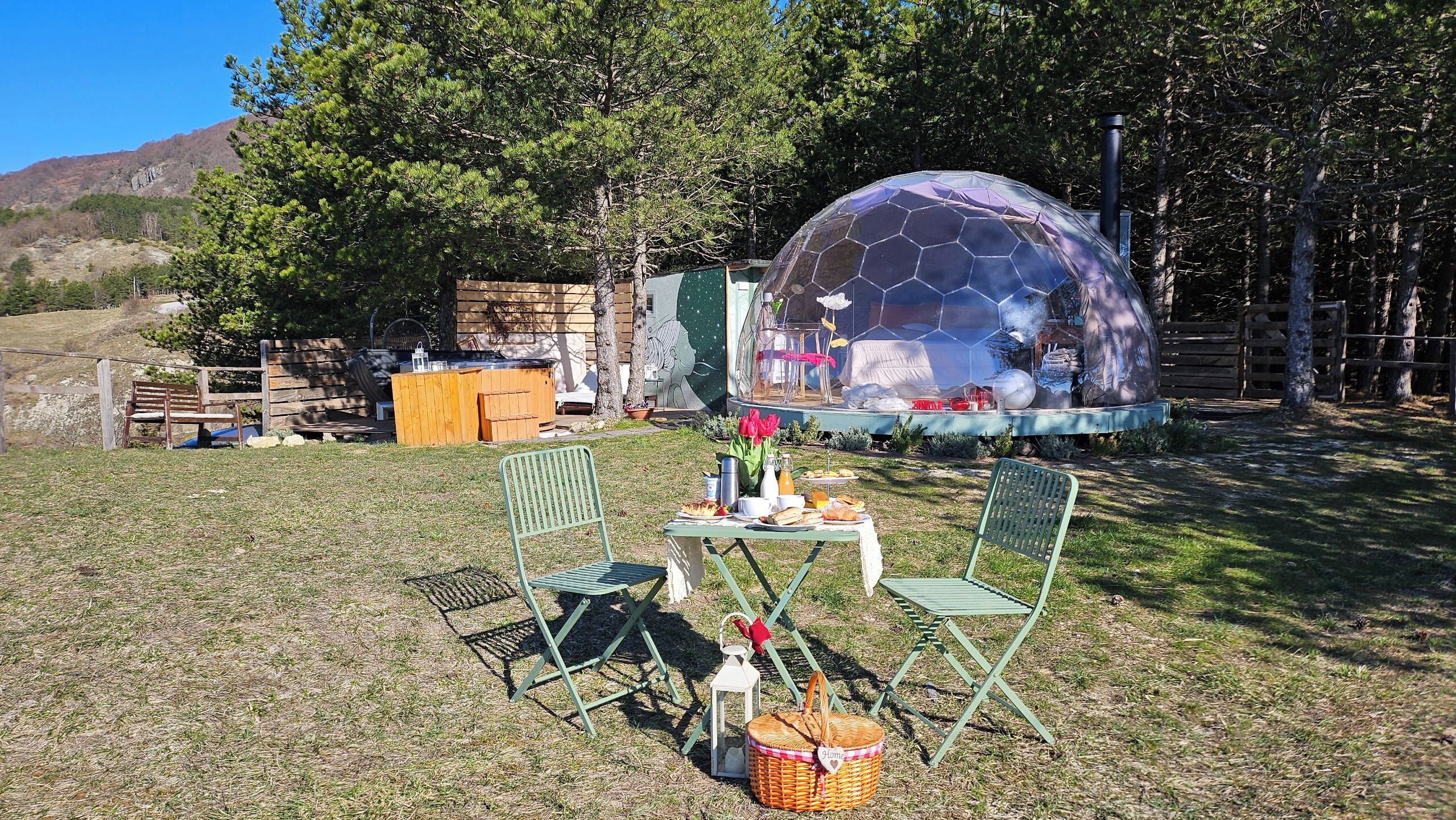 Picknickopstelling met een tafel, stoelen en een picknickmand in een grasrijke omgeving, met een geodetische koepelstructuur op de achtergrond.
