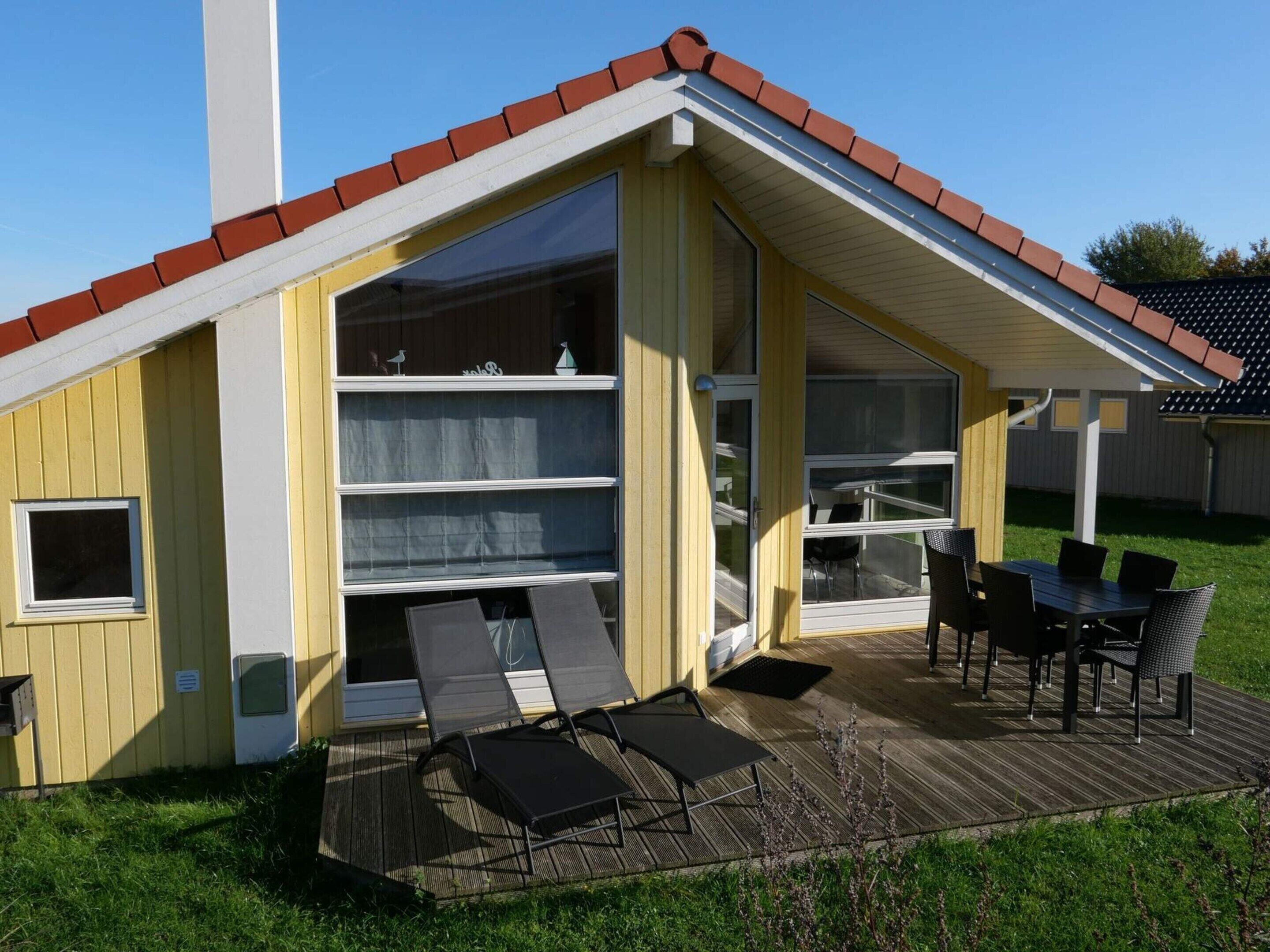 A small, single-story building with a sloped roof, large glass windows, and a deck with outdoor furniture.