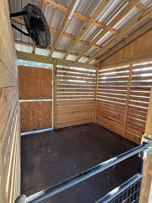 Un box pour chevaux équipé d’un ventilateur en métal, de lattes en bois et d’un tapis de sol noir.