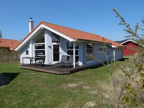 Ein eingeschossiges Haus mit rotem Ziegeldach, einer Veranda mit zwei Stühlen und einem sichtbaren Schornstein.