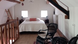 Ein Schlafzimmer mit Holzboden, einem Bett, zwei Nachttischen, einem Sessel, einer Kandelaber und zwei Fenstern.