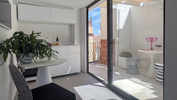 A modern kitchen with white cabinets, a dining table with a potted plant, and a glass door leading to an outdoor patio with a cactus and a pink candelabra. - Backhoe Suite (Taormina)