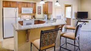 A kitchen with granite countertops, wooden cabinets, a built-in stove, a microwave, and a refrigerator. There are two bar stools and a television in the adjacent living area. - Wyndham Bonnet Creek, 3 bedroom deluxe suite, 3 BR/2 BA, kitchen, near EPCOT (Lake Buena Vista)