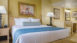 A hotel room with a bed, bedside tables, a nightstand, a lamp, a painting, a mirror, and a bathroom with a sink and toilet. - Wyndham Bonnet Creek, 3 bedroom deluxe suite, 3 BR/2 BA, kitchen, near EPCOT (Lake Buena Vista)