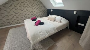 Ein Schlafzimmer mit schrägen Wänden, einem Oberlicht, einem Bett mit Kissen, einem Nachttisch und einer Wandleuchte.