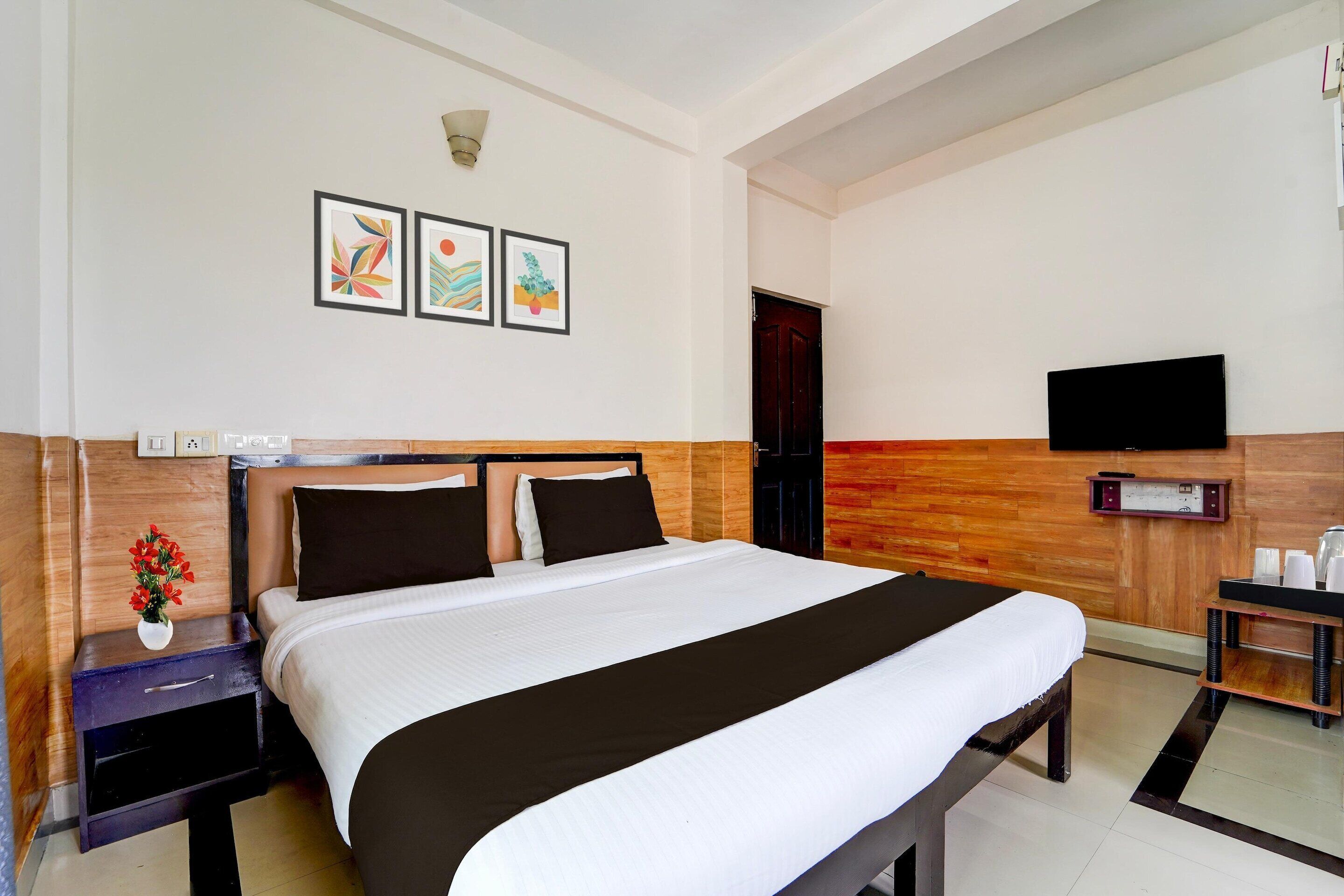 Ein Hotelzimmer mit einem Bett, einem Nachttisch mit Blumenvase, einem an der Wand befestigten Fernseher und Kunstwerken über dem Bett.