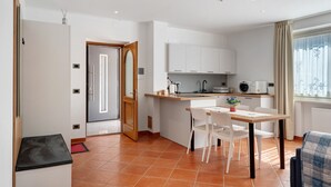 Een compacte keuken met witte kasten, een eethoek met een tafel en stoelen, en een raam met gordijnen.