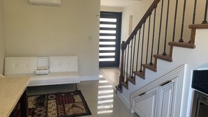 Uma sala de estar com sofá, mesa de centro, tapete estampado, escada com corrimão de madeira e ar condicionado fixado na parede.