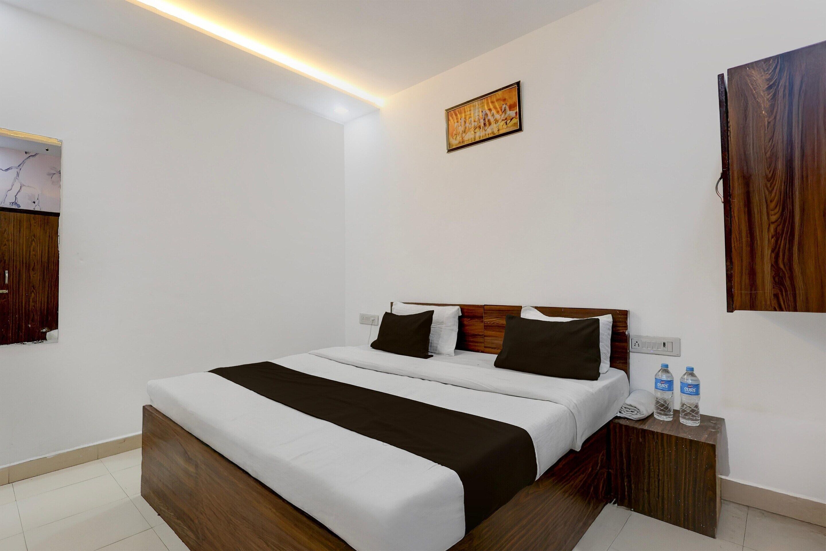 A hotel room with a wooden bed, white bedding, a nightstand with water bottles, and a painting on the wall.