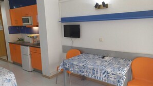 Una cocina compacta con microondas, refrigerador y gabinetes color naranja. Una mesa de comedor con mantel estampado y dos sillas color naranja se encuentran en el centro.
