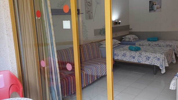 Una habitación con puerta de vidrio corredera, cama con colcha estampada, sofá con manta a rayas y una mesita con mantel de cuadros.